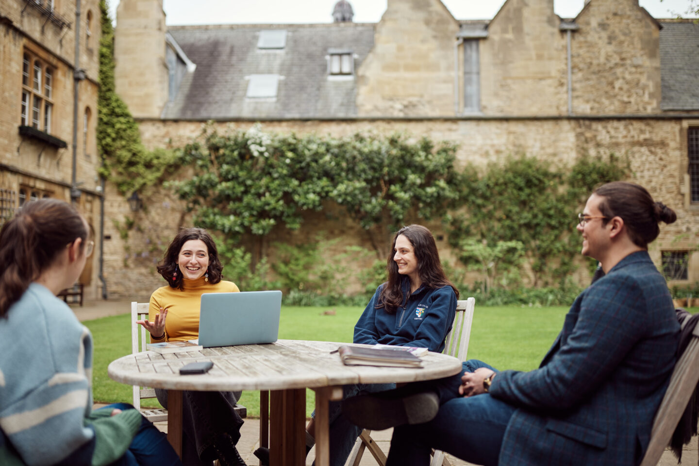 Photograph of four smiling students sitting at a table in a Lincoln College quad, a laptop open in front of one of them