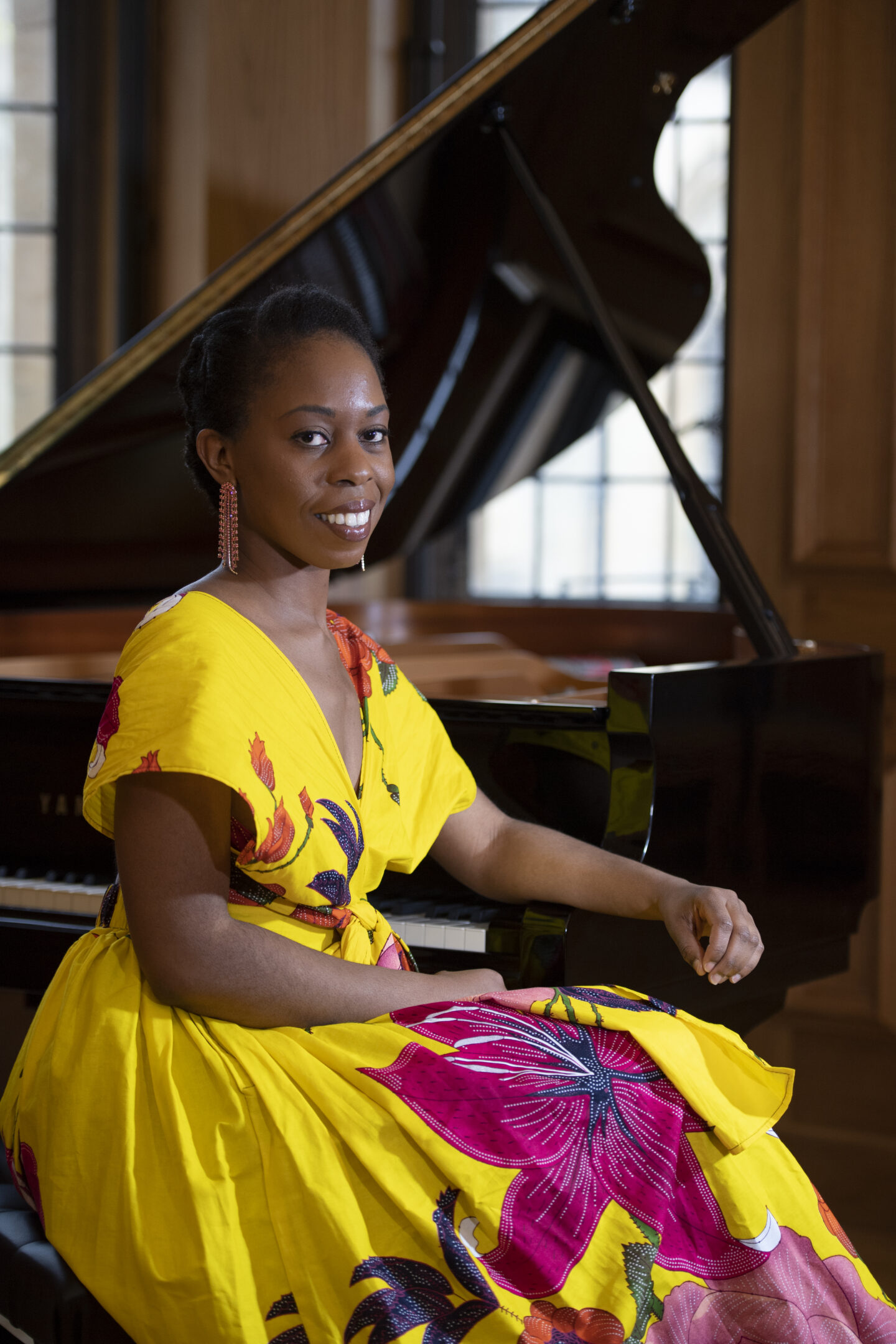 Headshot of Samantha Ege, a woman in a floral bright yellow dress, sitting with her arm rested against a piano