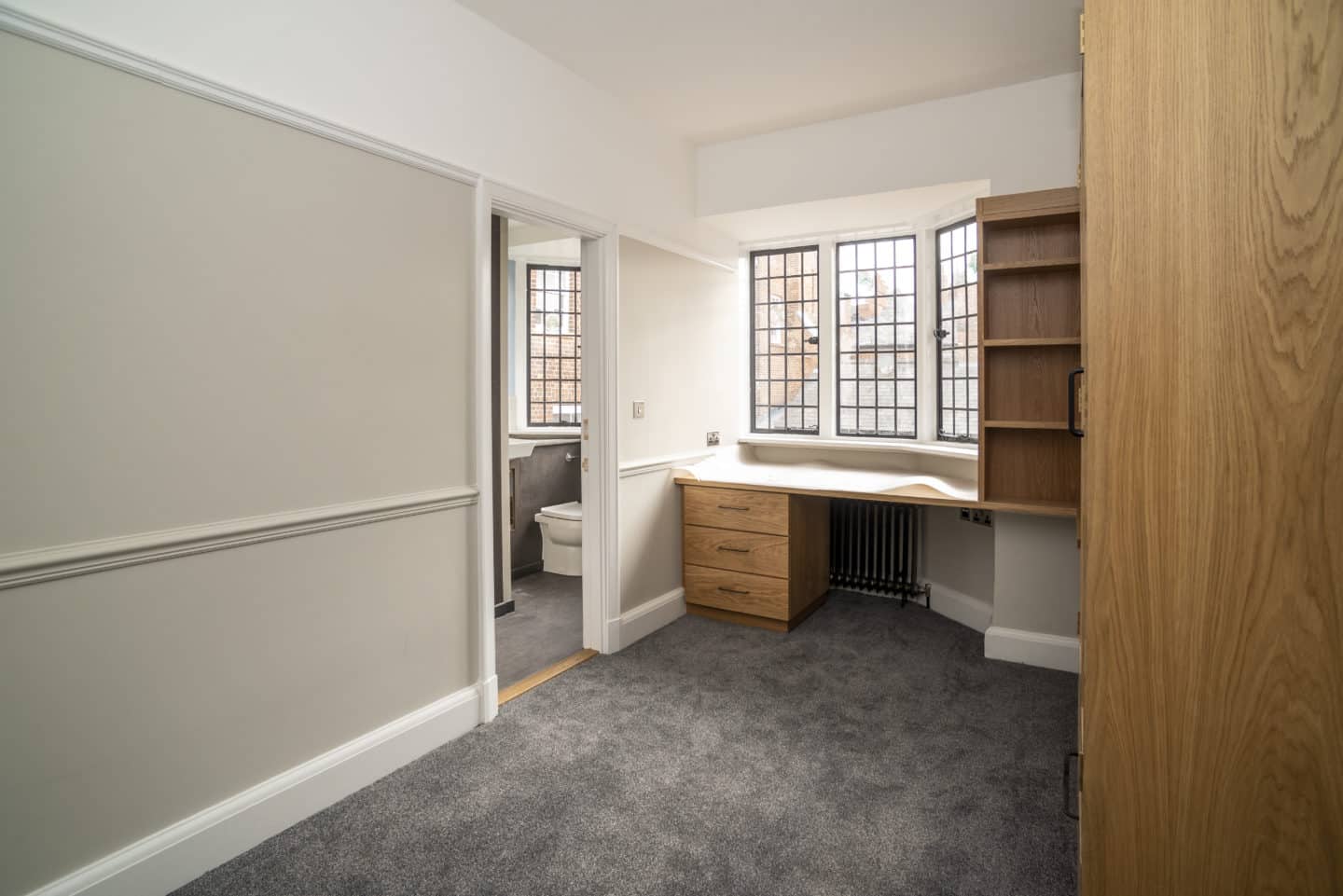 Part of a room with clean white walls, a desk with shelves and drawers, a radiator, windows, and an open door leading into a bathroom
