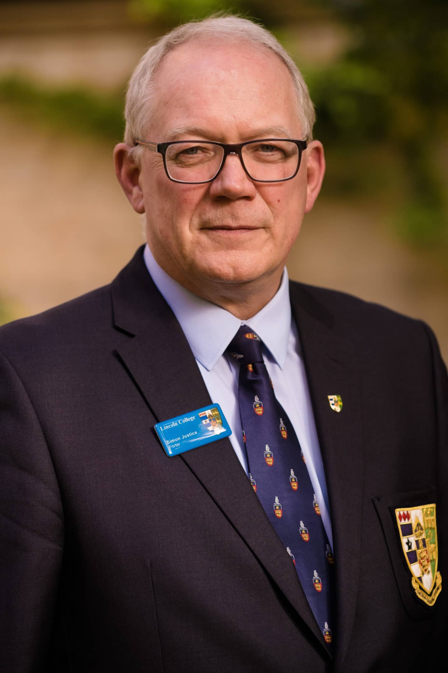 Headshot of Simon Justice, porter of Lincoln College wearing glasses, a suit with the Lincoln College crest, a name badge and Lincoln tie