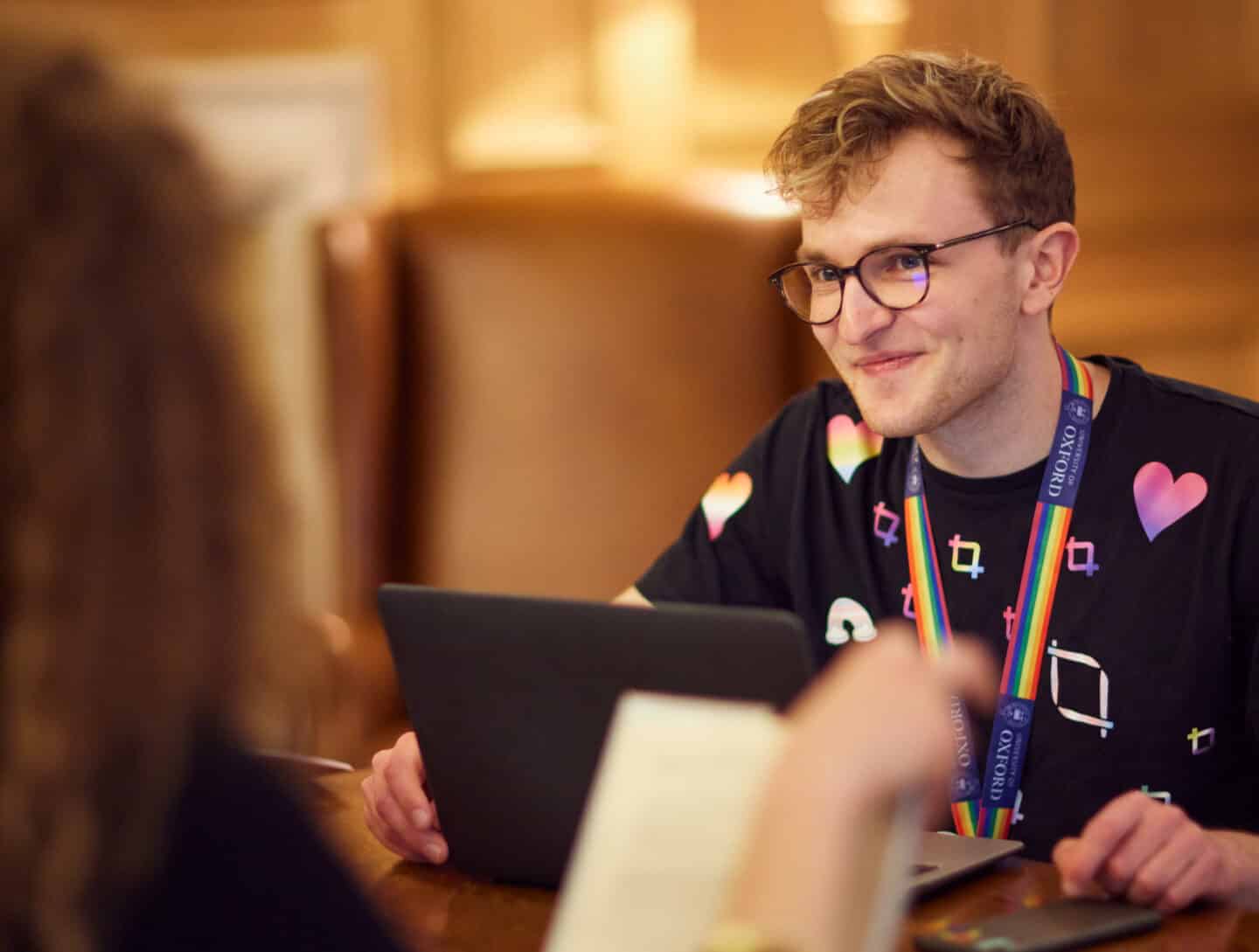 A student wearing glasses and a rainbow Oxford University lanyard, sitting at a desk with his laptop and watching someone else blurred in the foreground