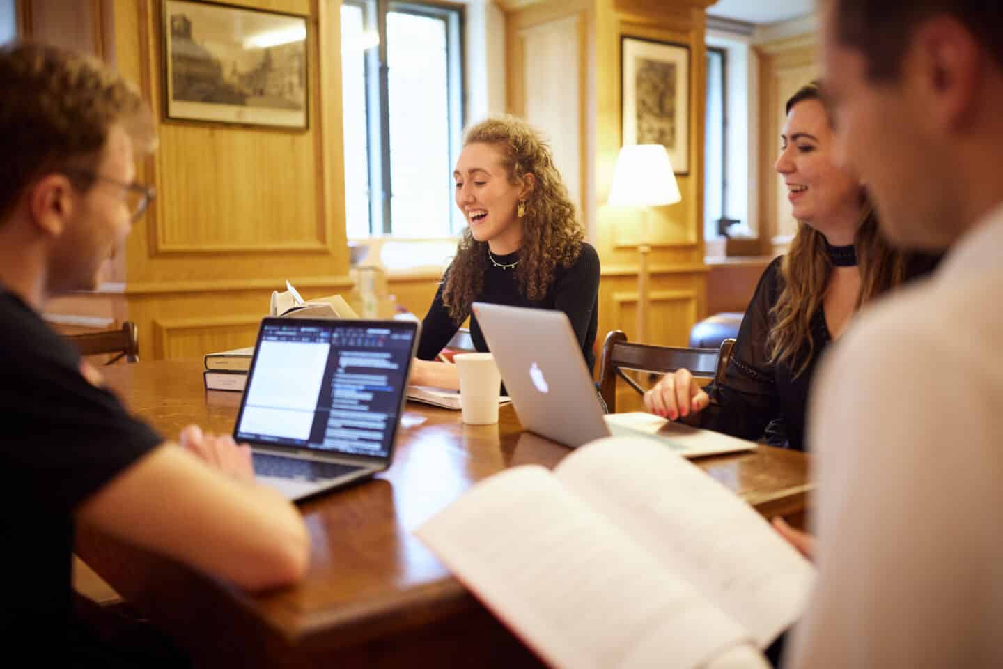 Four smiling students sitting around working with books and laptops at a wooden table in the Lincoln College Middle Common Room