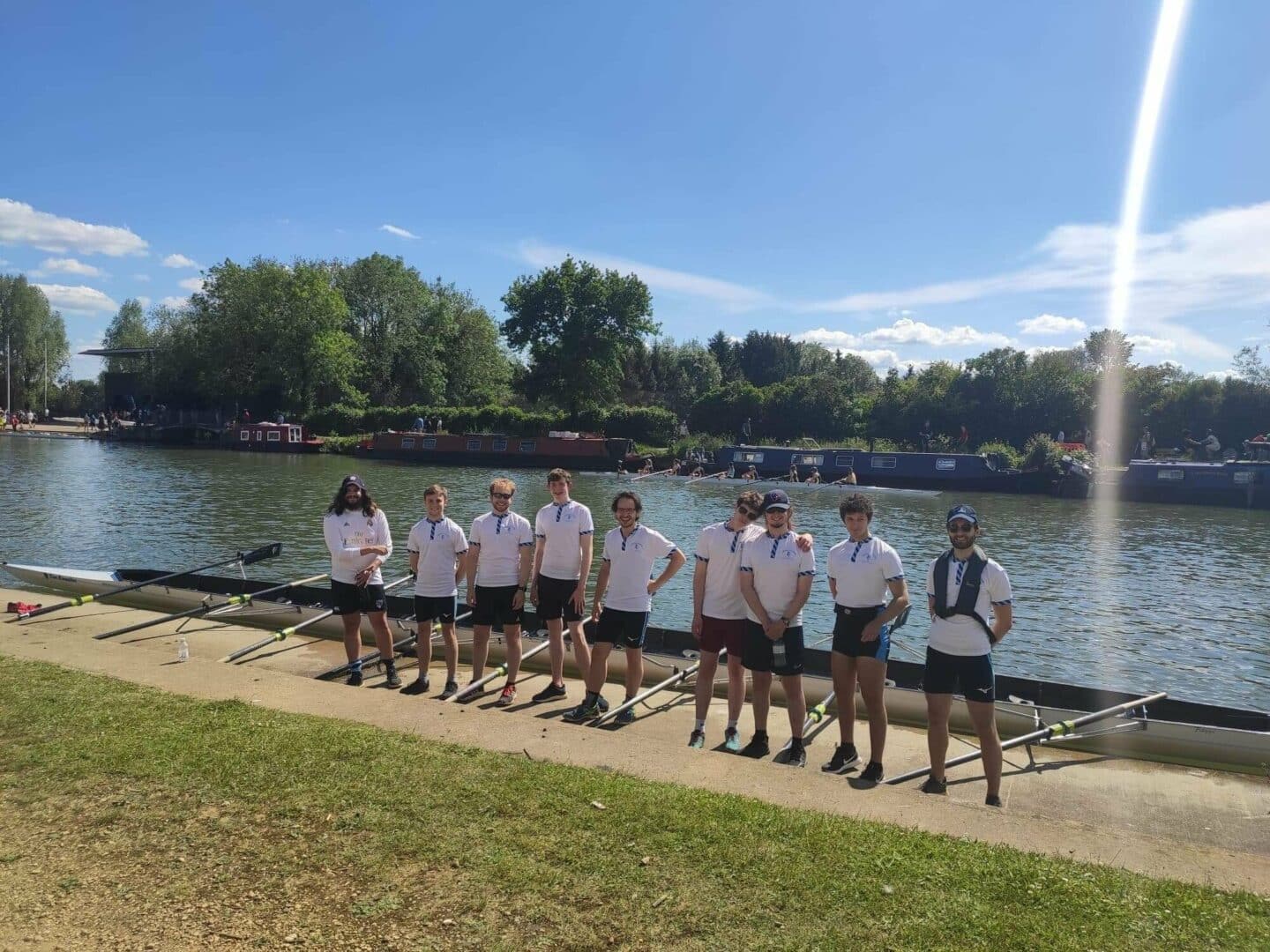Picture of the smiling Lincoln M2 boat club crew, all dressed in white t-shirts and shorts with their oars rested against their boat.