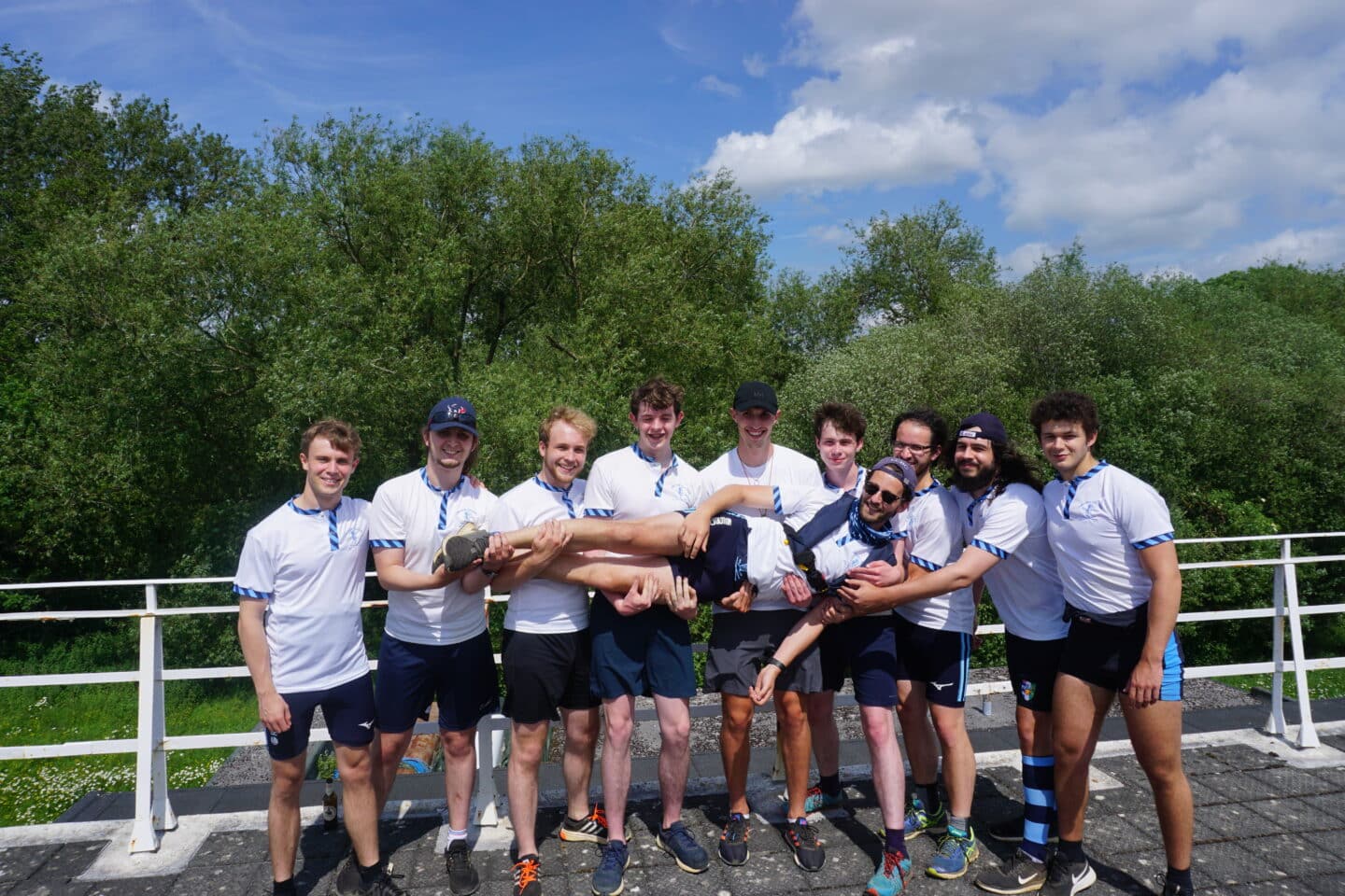 The M2 crew, all dressed in white t-shirts and shorts, smiling and holding up one member