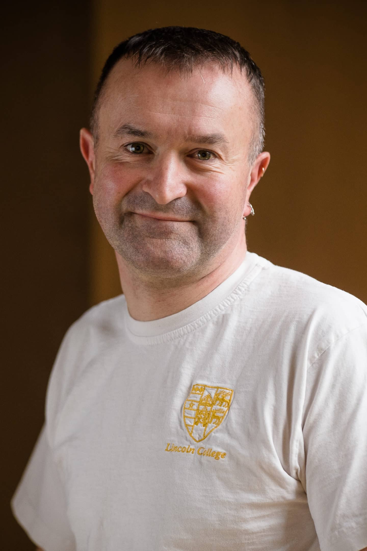 Headshot of Trevor Allan, a man in a white t-shirt with the Lincoln College crest in gold